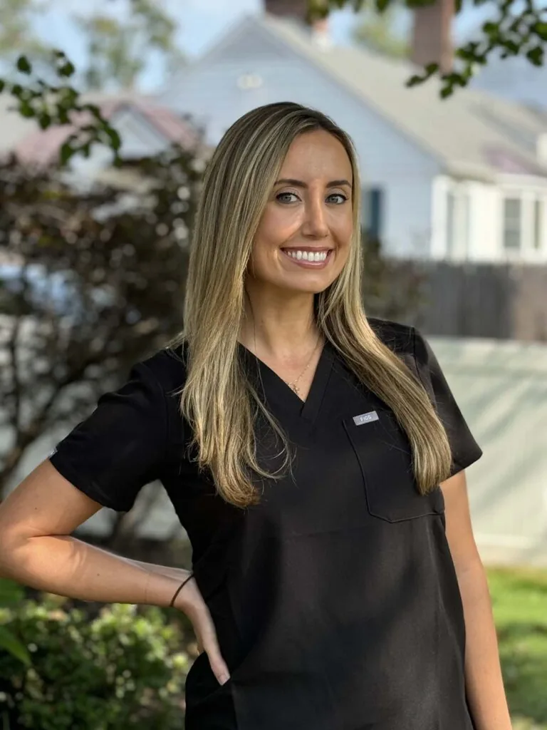 Dr. Amanda Baker, DMD, General Dentist at The Family Dental, is standing outdoors wearing black scrubs. The background features a house and some trees, creating a natural and professional setting.
