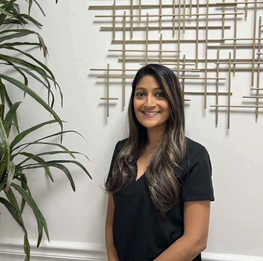 Dr. Amie Shah, DMD, Pediatric Dentist at The Family Dental, is standing in front of a white wall with a decorative metal grid pattern. She is wearing a black top and has long, wavy hair. There is a large green plant to her left.