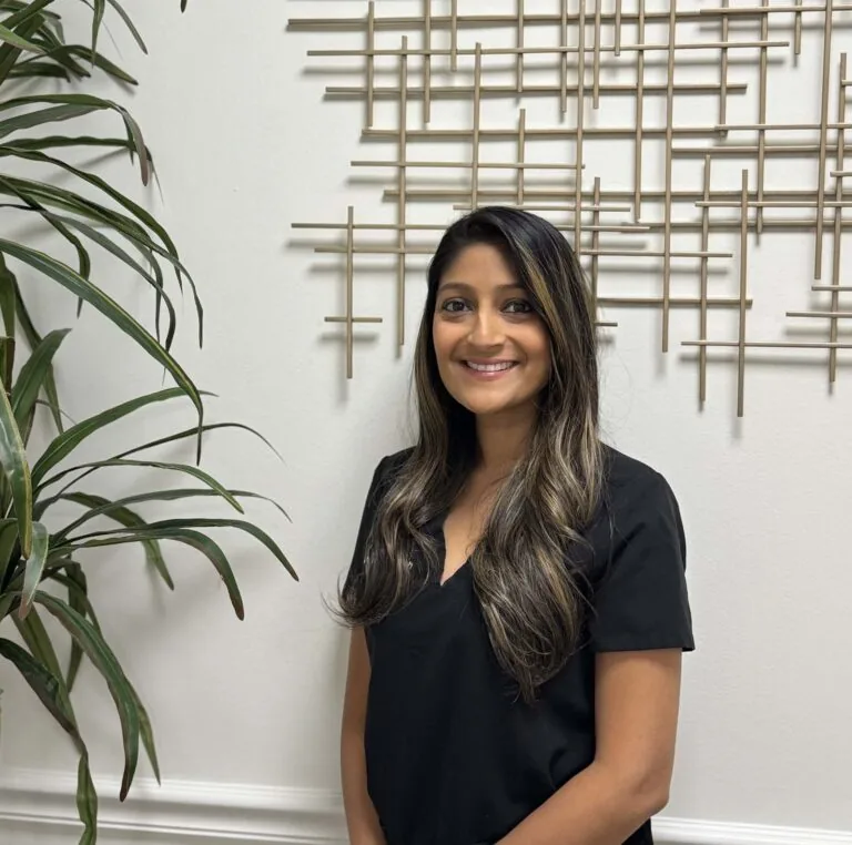Dr. Amie Shah, DMD, Pediatric Dentist at The Family Dental, is standing in front of a white wall with a decorative metal grid pattern. She is wearing a black top and has long, wavy hair. There is a large green plant to her left.