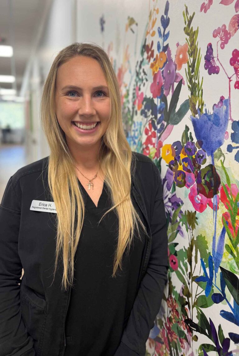 Erica, a Dental Hygienist at The Family Dental, is standing in front of a colorful floral mural. She is wearing a black jacket with a name tag.