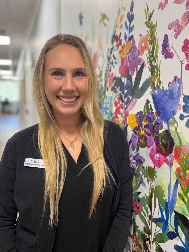 Erica, a Dental Hygienist at The Family Dental, is standing in front of a colorful floral mural. She is wearing a black jacket with a name tag.