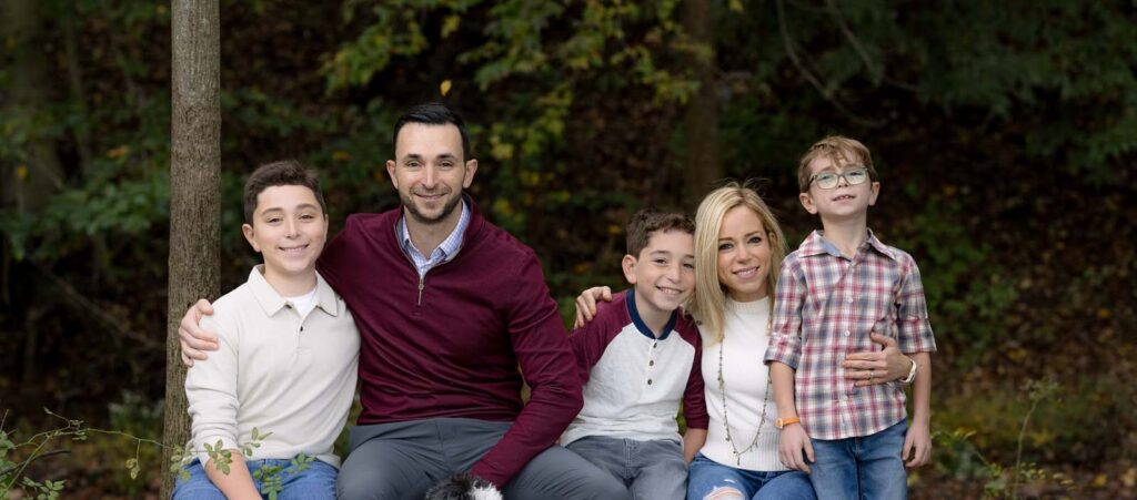 Dr. Brett Druger is sitting outdoors in a wooded area with his wife and their three kids. Dr. Druger is wearing a maroon sweater, and his wife is wearing a white shirt. Their three children are dressed in casual clothing: one in a white and maroon shirt, one in a plaid shirt, and one in a white shirt. The family is surrounded by trees and foliage, indicating a natural setting.