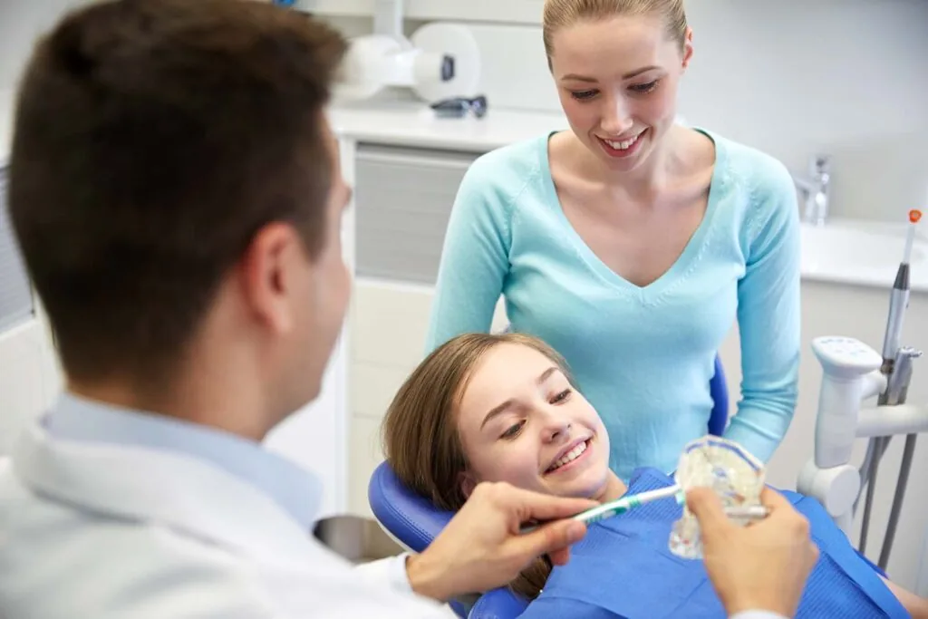 people, medicine, stomatology and health care concept - happy male dentist with toothbrush and jaw layout showing how to brush teeth to patient girl and her mother at dental clinic office